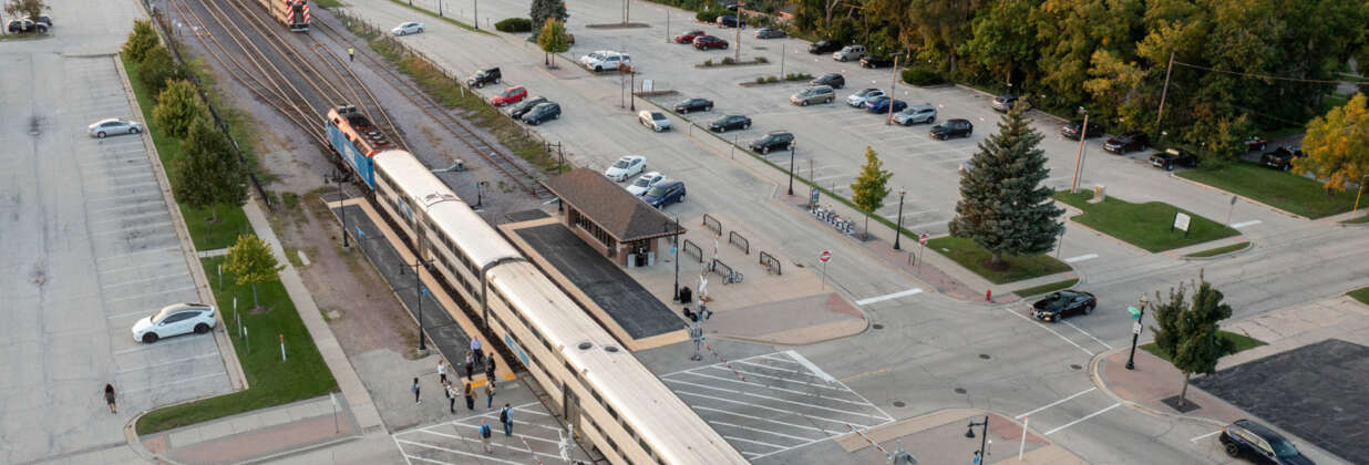 A Metra train at a station with a half-full parking lot and pedestrians exiting the train.