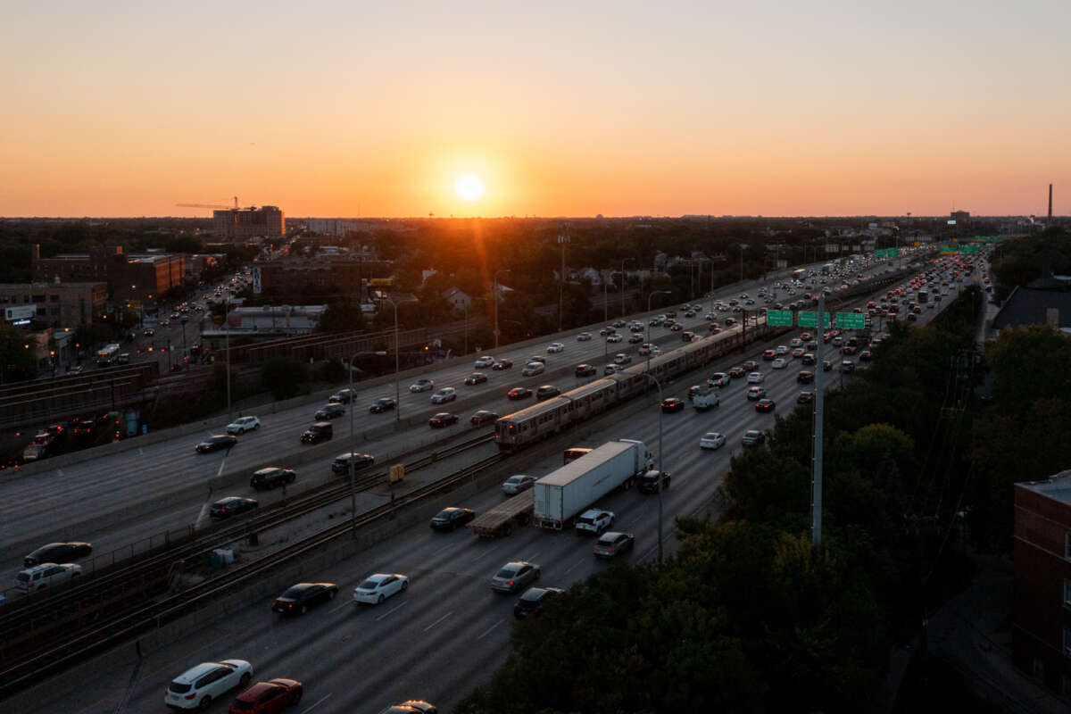 Wide shot of a CTA train running along the middle of a highway with the sun near the horizon in the background.