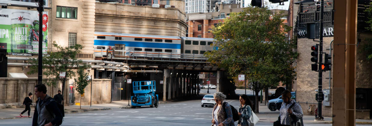 People with backpacks cross a street in downtown Chicago as an elevated Metra train goes by in the background.