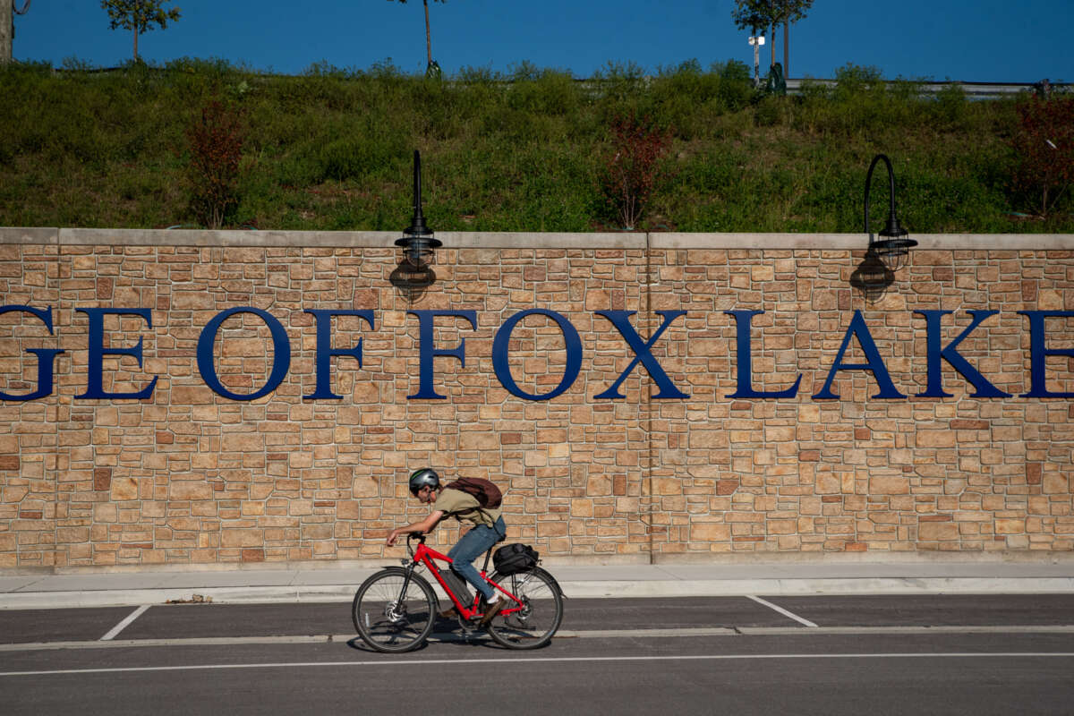A cyclist on an electric bicycle rides past a brick wall with a "Village of Fox Lake" sign on it.