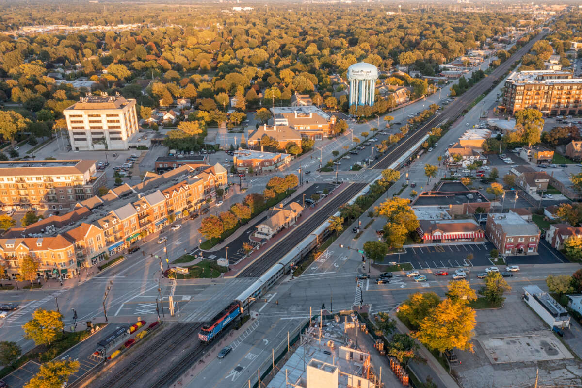 A wide shot of a Metra train at a suburban station with dense development across the street.