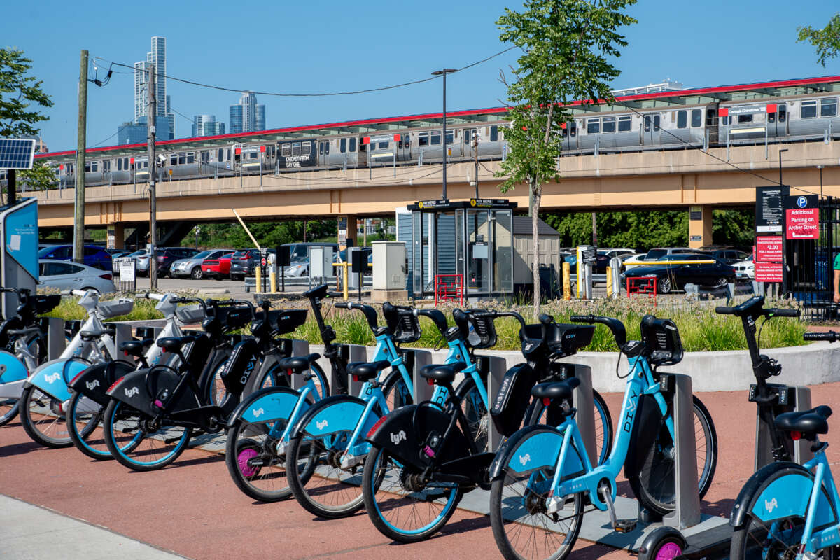 A Divvy bikeshare rack full of bikes with a CTA train at the Cermak Chinatown station and skyscrapers in the background