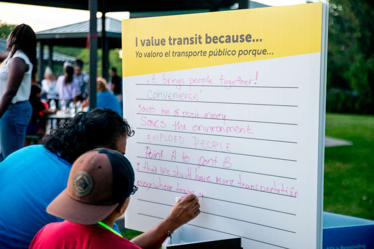 An adult with a child writes on a sign with the prompt, "I value transit because..."