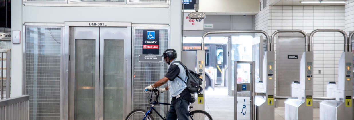 A man with a bicycle walks through the accessible turnstile toward an elevator at a CTA station.
